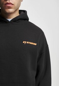 Black hooded sweatshirt made of cotton, featuring a small orange graphic logo "2Y STUDIOS" on the chest. Soft texture, loose fit.