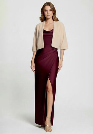 Femme portant une longue robe bordeaux avec une fente sur la cuisse et un cardigan court beige, debout devant un fond clair uni.