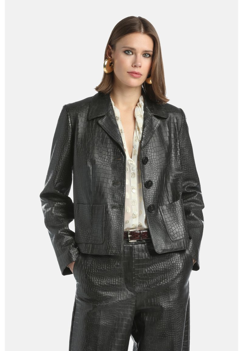 Vrouw met schouderlang bruin haar, gekleed in een zwarte leren jas met krokodillenpatroon en bijpassende broek, een witte blouse met een patroon en grote gouden creolen.
