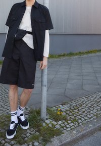 Chemise noire à manches courtes sur un haut en tricot blanc, associée à un short noir. Rayures blanches remarquables sur les chaussettes et baskets noires.