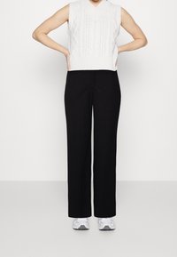 Personne portant un pull sans manches en tricot blanc, un pantalon noir ample et des baskets blanches, debout avec les mains sur les hanches.
