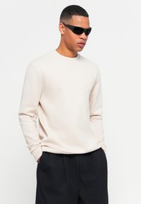 Sudadera de cuello redondo color beige claro con mangas largas y un detalle sutil del logo. Gafas de sol negras, combinadas con pantalones oscuros, completan el conjunto.