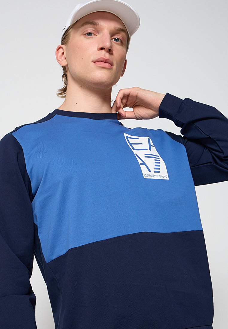 Suéter azul y marino con mangas largas, que presenta un parche con logo y una tela texturizada. El diseño alterna colores horizontalmente a lo largo del pecho.