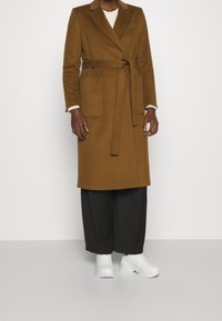 Cappotto marrone in misto lana con vita stretta da cintura, colletto a rever e due tasche frontali, abbinato a pantaloni neri larghi e sneakers bianche.