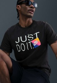 Camiseta de algodón negra con el texto "JUST DO IT" en blanco y un logotipo de Nike colorido en una etiqueta cuadrada. Combinada con pantalones cortos oscuros y gafas de sol.