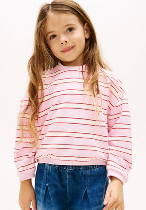 Νεαρή κοπέλα με μακριά μαλλιά που φοράει ροζ φούτερ με λεπτές κόκκινες ρίγες και μπλε τζιν παντελόνι, στέκεται μπροστά σε ένα απλό φωτεινό φόντο.