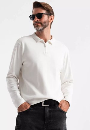 Hvid langærmet polo shirt med en halvdelsk ziplukning, båret med sorte slidte jeans og et sort bælte, kombineret med mørke solbriller.