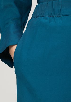 Tyrkysová tkanina s hladkou texturou má nabráný pas. Ruka je viditelná v boční kapse oděvu.