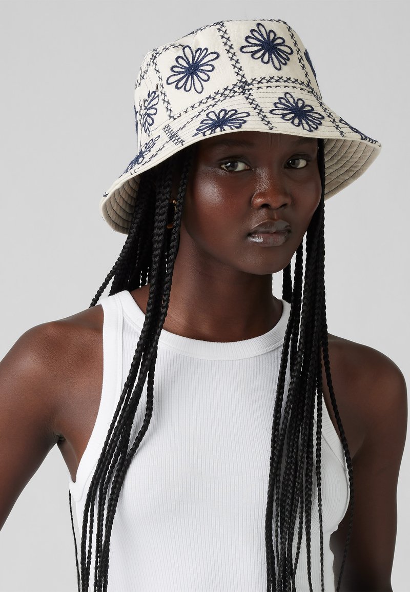 Cappello a secchio in tessuto crema con ricami floreali navy e motivi cuciti; una visiera larga e morbida e una superficie testurizzata.