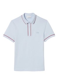 Polo Lacoste bleu clair à manches courtes, avec un liseré rouge sur le col, la patte de boutonnage et les poignets, et un petit logo crocodile sur la poitrine.