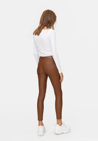 Leggings en simili cuir marron avec une taille haute, coupe slim et poches arrière, associés à un haut blanc à manches longues et des baskets blanches.
