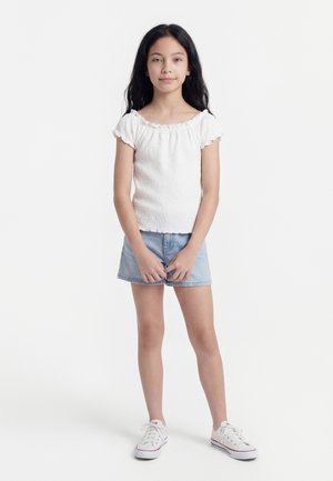 Νεαρή κοπέλα με μακριά σκούρα μαλλιά στέκεται κατά πρόσωπο, φορώντας λευκό κοντομάνικο τοπ, ανοιχτόχρωμα τζιν σορτς και λευκά αθλητικά παπούτσια.