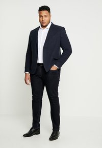 Marineblazer met twee zakken, wit overhemd en donkere jeans. Gelaagd stof, slim fit en zwarte schoenen maken de look compleet.