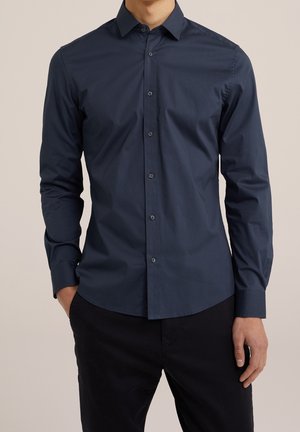 Mann trägt ein tailliertes, dunkelblaues Hemd mit langen Ärmeln, das über schwarze Hosen untendrunter getragen wird, steht mit einer Hand in der Tasche.