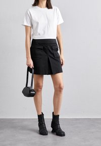 Mujer vestida con una camisa blanca de manga corta, falda negra con bolsillos frontales, botas negras hasta el tobillo, sosteniendo un pequeño bolso negro a su lado.