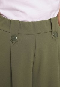Pantalon vert olive en tissu lisse, avec une taille haute, deux boutons en guise d'accent, et de légers plis pour une silhouette ajustée.