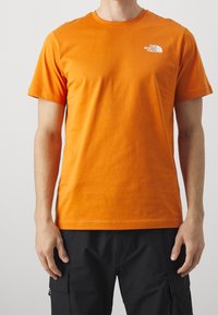 Oranžové bavlněné tričko s kulatým výstřihem a krátkým rukávem. Na levé části hrudi se nachází malé bílé logo. Hladký, jemný povrch.