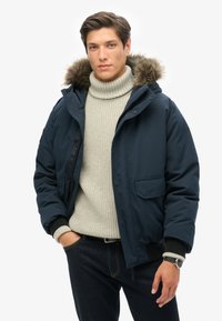 Marineblå polstret jakke med en hætte foret med pels, med en frontlynlås, ribbede manchetter og en brystlomme. Båret over en lys grå sweater.