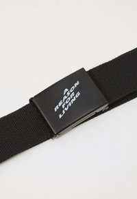 Zwarte stoffen riem met een rechthoekige metalen gesp waarop in witte tekst staat: "EEN REDEN OM TE LEVEN." De tekst is schuin geplaatst voor extra nadruk.