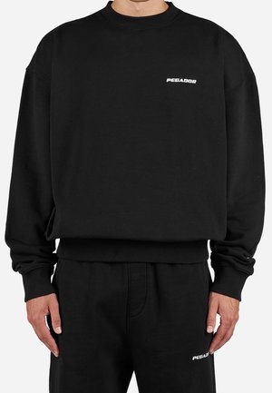 Mann trägt einen schwarzen Pullover und passende Hose, beide mit kleinen weißen "PEGADOR"-Logos auf der Brust und dem Oberschenkel.