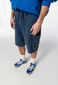 Shorts bleu marine avec une poche et des détails de marque, associés à des baskets bleues, grises et jaunes. Le modèle porte des chaussettes blanches.