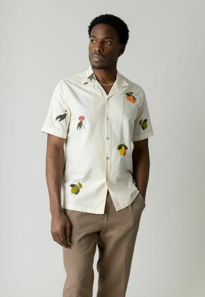 Mand iført en off-white kortærmet skjorte med frugt- og blomstermønster, beige bukser, stående med den ene hånd i lommen mod en ensfarvet baggrund.