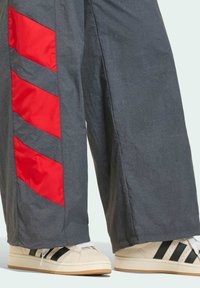 Sive široke hlače imaju crvene dijagonalne pruge sa strane, u paru s kremastim tenisicama koje imaju crne naglaske i bijele vezice.