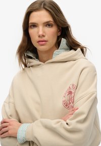 Beżowa bluza z kapturem z przednią kieszenią kangurką i różowym logo graficznym. Warstwowa z pasiastą niebiesko-zieloną koszulą przy kołnierzu. Miękka tkanina.