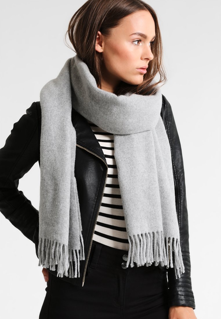 Lys grå uldscarf med frynsede ender, viklet rundt om halsen og parret med en sort læderjakke og en stribet bluse.