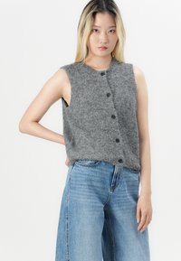 Joven mujer con cabello largo y rubio lleva un suéter gris sin mangas abotonado y pantalones vaqueros de pierna ancha, de pie con una mano en la cadera.