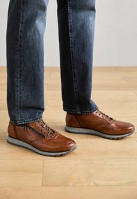 Scarpe in pelle marrone con finitura texture, suola in gomma grigia e design con lacci, dotate di una cerniera laterale e branding minimalista.