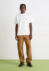 Tricou alb cu mânecă scurtă, dotat cu un buzunar pe piept și un logo cu inimă roșie, asociat cu pantaloni de culoare maro deschis, drepți, și pantofi negri.