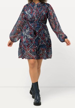 Femme portant une robe à manches longues, longueur genou, avec un motif cachemire bleu, rouge et blanc et des bottines noires.