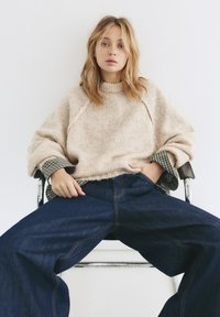 Béžový pletený sveter s vysokým golierom a spustenými ramenami, prehodený cez zelenú kockovanú košeľu, v kombinácii s širokými tmavomodrými džínsami.