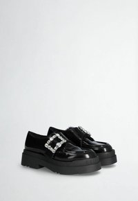 Black patent leather shoes with a thick black sole and silver buckle accent. Square shape, smooth texture, and rounded toe design.