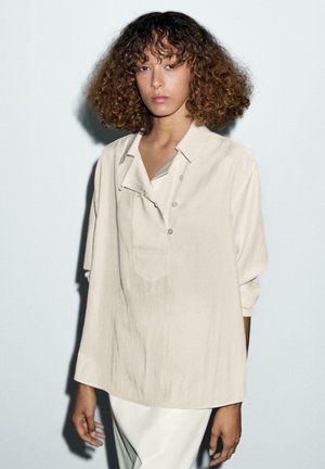 Chemise oversized beige clair à manches courtes, avec une patte de boutonnage, une poche poitrine unique et un tissu texturé.