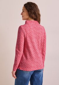 Röd topp i slubtextur med hög krage, med diagonala vita ränder, långa ärmar och sidofickor, i kombination med blåjeans.