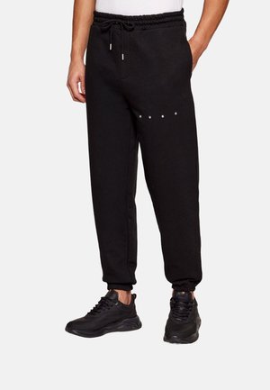 Hombre vistiendo pantalones de chándal negros con cintura y puños elásticos, logo blanco en el muslo izquierdo, combinados con zapatillas negras y camisa blanca.