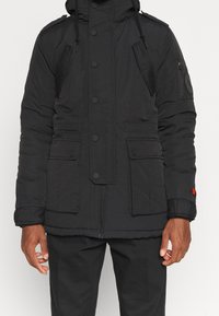 Veste rembourrée noire avec capuche, dotée de boutonnage, de multiples poches et d'un tissu texturé. Comprend des patchs décoratifs sur les manches.