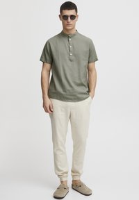 Maslinasto zeleni kratki rukav Henley majica s tri gumba, kremaste uske hlače i bež slip-on cipele. Glatka tekstura, opušteni kroj.