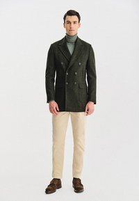 Ciemnozielony wełniany płaszcz dwurzędowy z szarymi guzikami, noszony na jasnozielonym golfie, w połączeniu z kremowymi spodniami i brązowymi mokasynami.