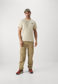 Beige, rövidujjú póló fekete logóval, khaki cargo nadrág cipzáras zsebekkel és piros túracipő. Világoszöld sapka.