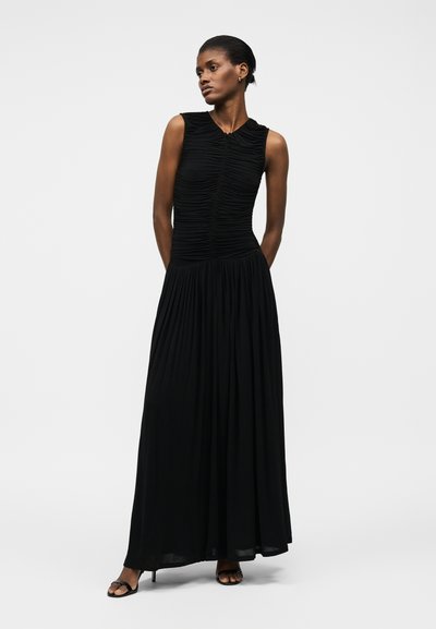 Naine mustas varrukateta maxi-kleidis, millel on voltidega bodi ja volditud seelik, seisab käed selja taga lihtsa tausta ees.