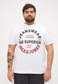 Valge puuvillane T-särk, millel on julge punane ja tume sinine graafiline print, sealhulgas tekstid "JEANSWEAR" ja "JACK & JONES."