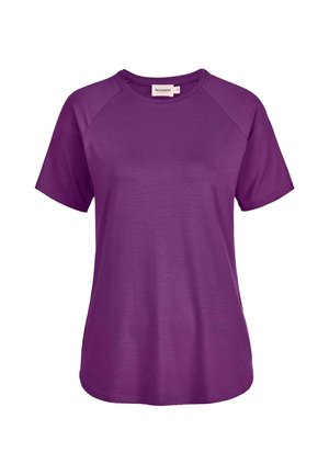 Junge Frau mit glatten braunen Haaren, die ein schlichtes lila Kurzarmshirt und schwarze Hosen trägt, stehend mit den Händen nahe der Hüften.