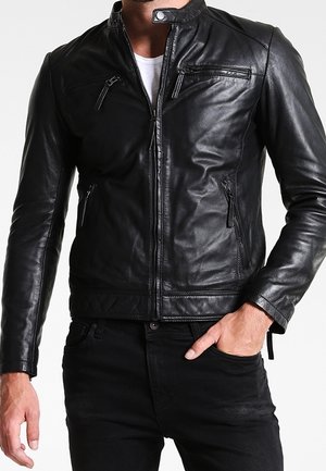 Homme portant une veste en cuir noire par-dessus une chemise blanche, une main dans la poche d’un pantalon noir, sur un fond uni.