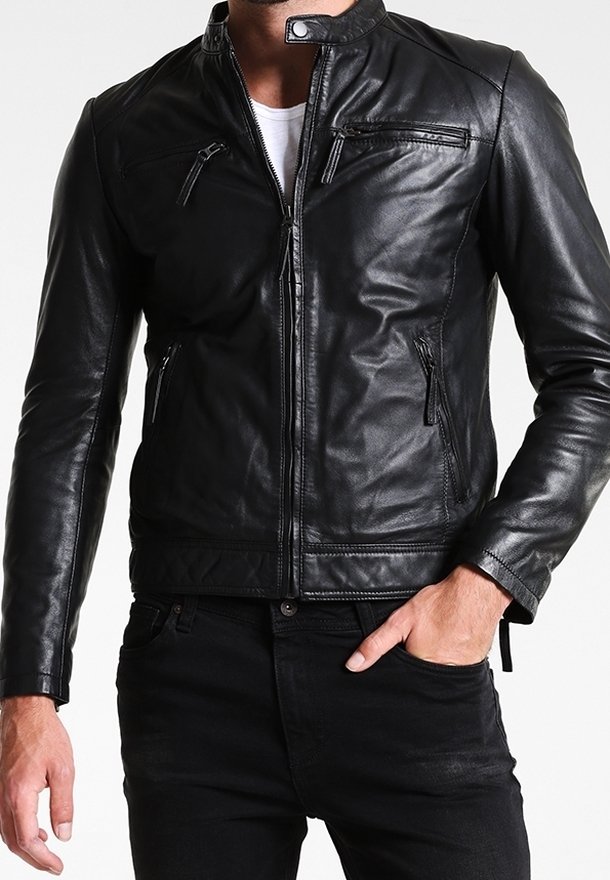 Homme portant une veste en cuir noire par-dessus une chemise blanche, une main dans la poche d’un pantalon noir, sur un fond uni.