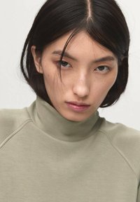 Suéter de cuello alto ajustado en verde oliva claro, con una textura suave, detalles de costura y un cuello alto. El modelo tiene el cabello oscuro y liso.