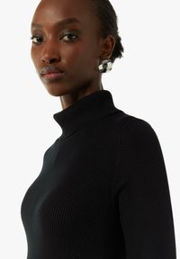 Schwarzer, gerippter Rollkragenpullover mit tailliertem Design. Verfügt über einen hohen Kragen und strukturierte Strickware. Silberne Statement-Ohrringe runden den Look ab.