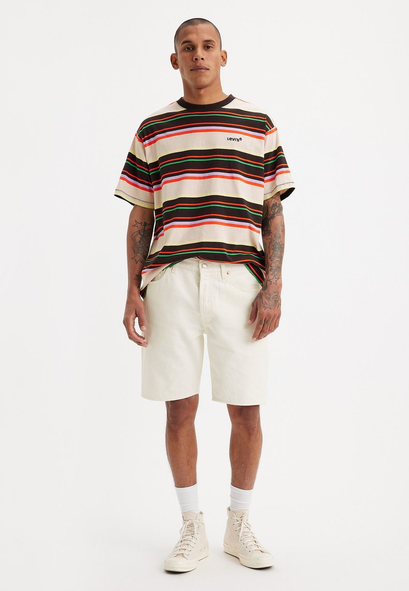 Gestreiftes T-Shirt mit kurzen Ärmeln in Creme, Braun, Grün und Rot; kombiniert mit weißen Denim-Shorts. Das Model trägt beigefarbene High-Top-Sneaker.